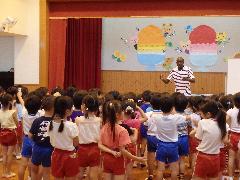 かき氷の壁面飾りがされている部屋で正面に立って話をしているALTと、話を聞いている園児たちの写真