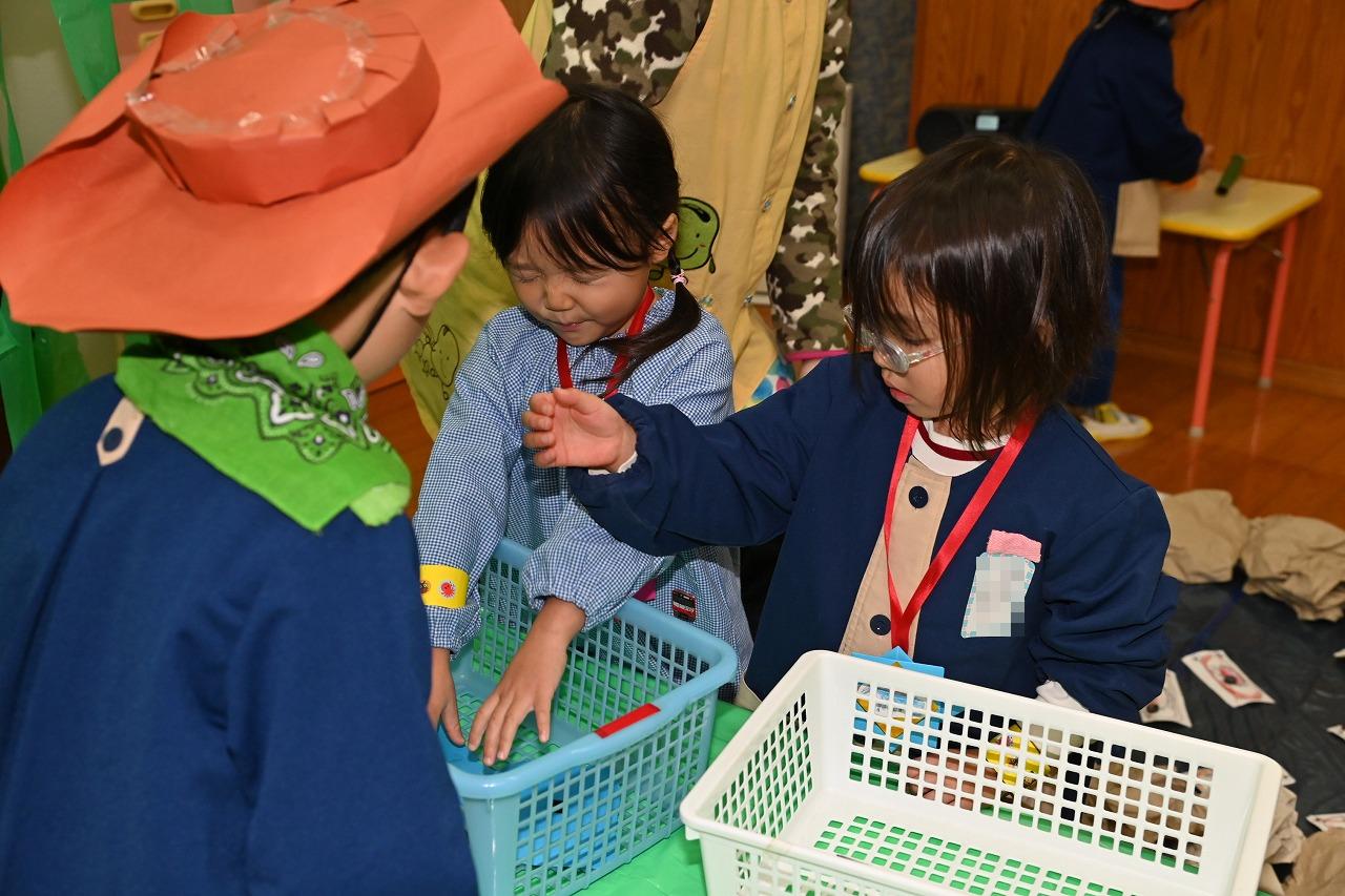 年少児が目を閉じて、かごの中に入れてあるカードを選んでいる。その隣に女児1名、前に年長児が立って見守っている様子