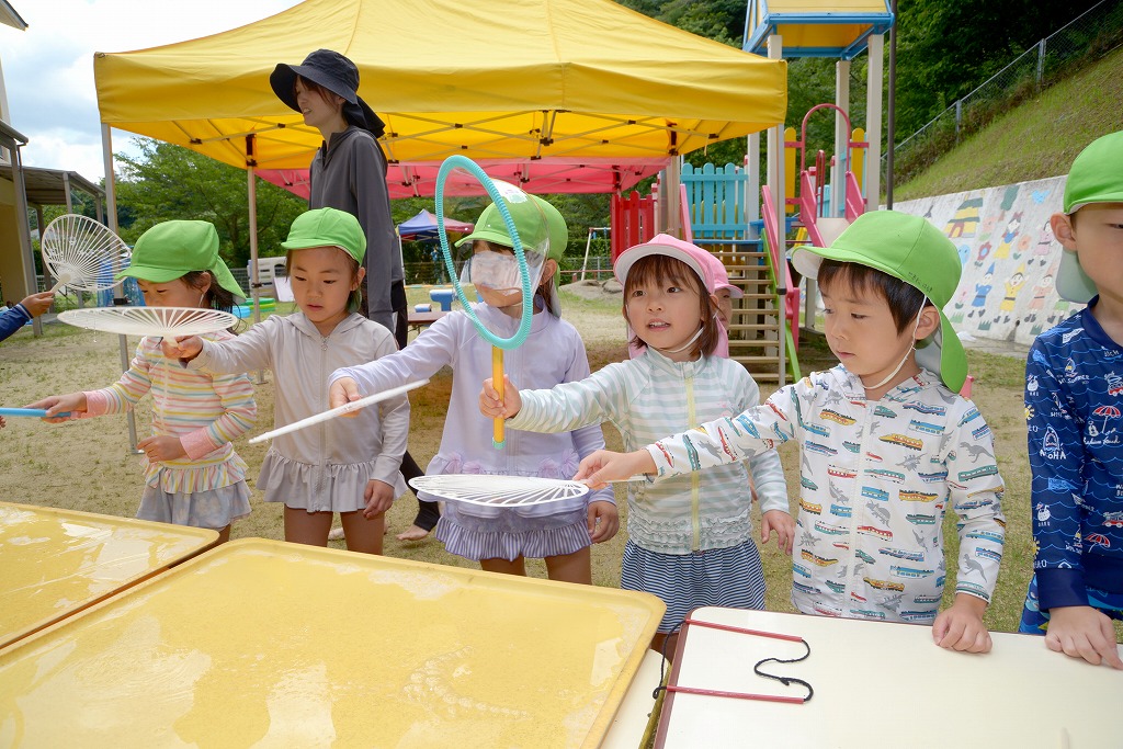 園児たちが、うちわの骨組やプラスチックの輪っかなど、好きなものを持ってシャボン玉を作ろうとしている様子