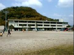 後方に山があり、3階建ての校舎の前には広々とした校庭が広がり、児童達が活動をしている立花小学校の外観写真