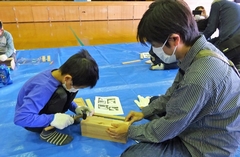 ブルーシートの上で、お父さんが木材をおさえ、男の子がくぎを打っている様子の写真