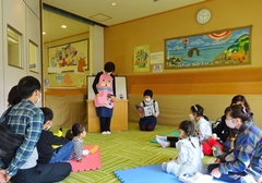 読み聞かせをしている女性の方とぬいぐるみを持って聞いている子供たちと保護者の方々の写真