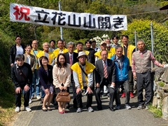立花山山開きと書かれた旗の下に、黄色いベストを着た関係者や町長などが並んでいる記念写真