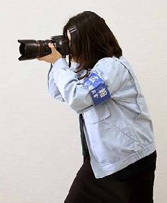 グレーの上着に腕章を付けた女性がカメラを構えている様子の写真