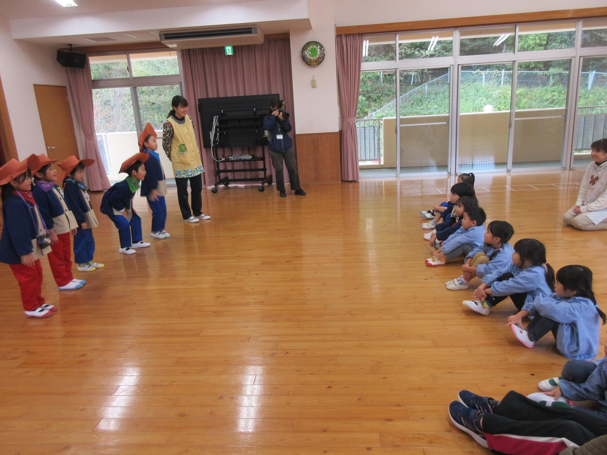 遊戯室で、年長児5名と年中、年少児が向き合って、年長の話を聞いている様子