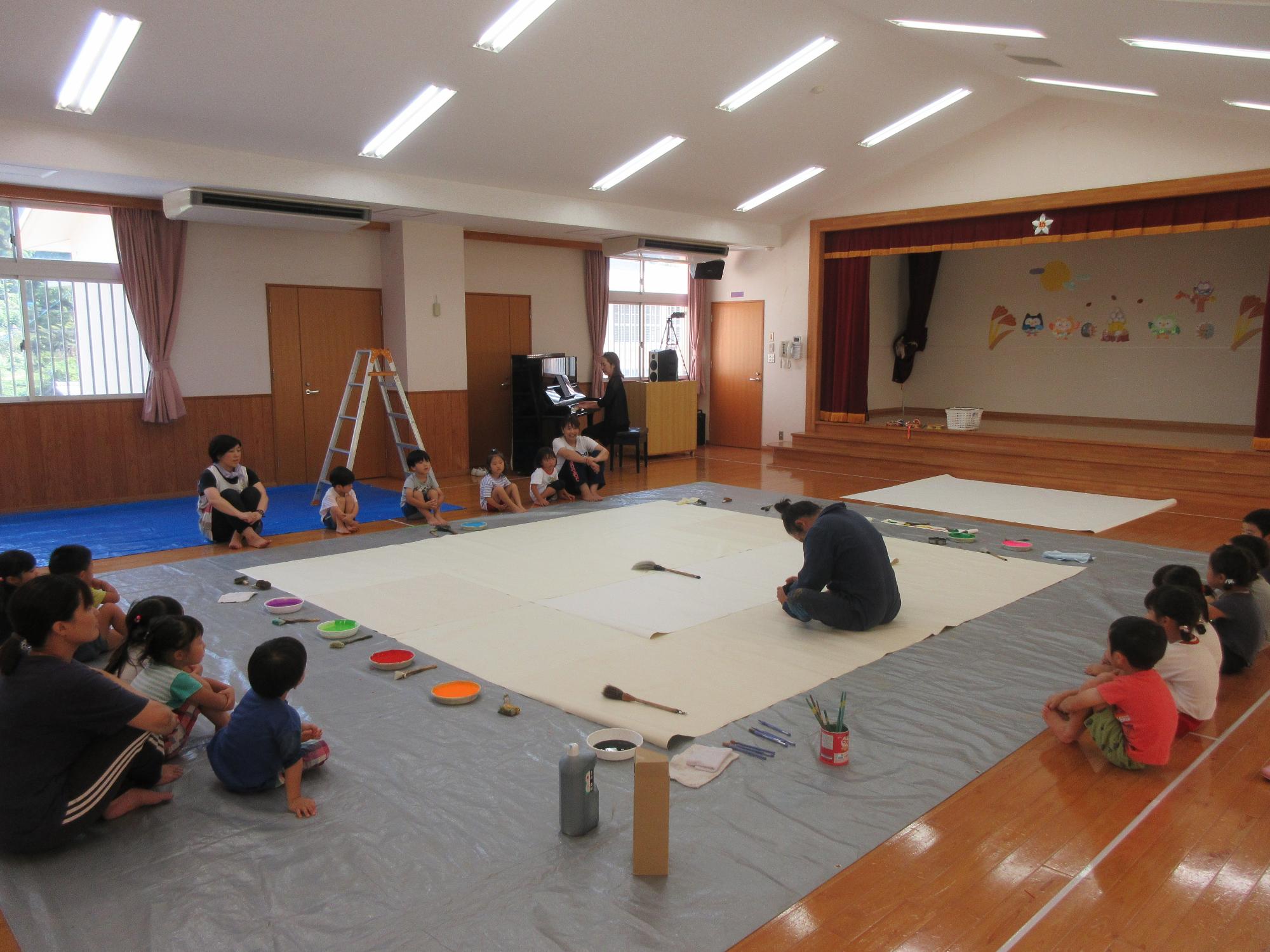 三四郎さんが大きな紙の上で集中力を高めているのを、まわりから子どもたちや先生が見守っている様子
