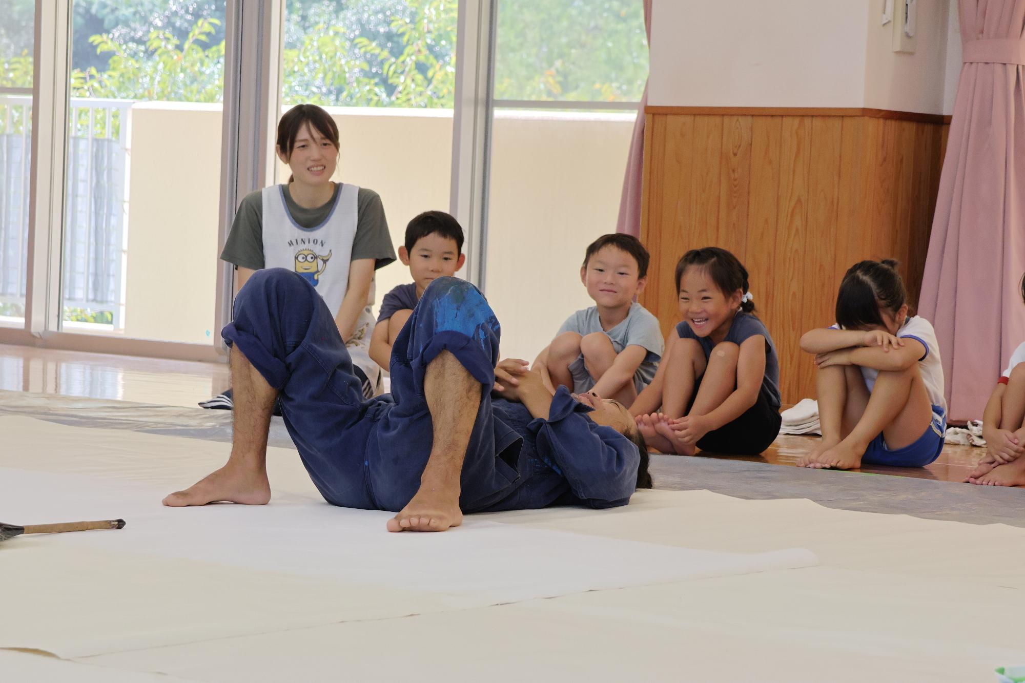三四郎さんが大きな紙の上にあおむけに寝ている。その様子を年長児が得外で見ている様子。