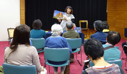 紅葉の木が書かれた絵本の見開きページを開き、参加者の大人たちに読み聞かせをしている女性の写真