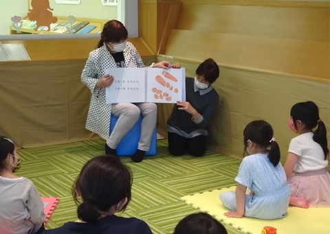 椅子に座った女性が絵本を膝の上に乗せて持ち、床に座った女性が見開きページを持ち、子供たちに読み聞かせをしている様子の写真