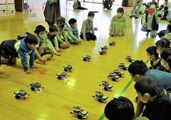 子供たちが向かい合うように2列に座り、同じく子供たちのロボットが向かい合うように2列で並んでいる写真