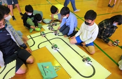 床に敷かれたマットに書かれたレーンの上を進むロボットとそれを見ている子供たちの写真