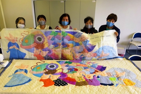 5人の女性がカラフルなうろこの魚とこぶたのイラストが描かれた大きな布のタペストリーを持っている写真