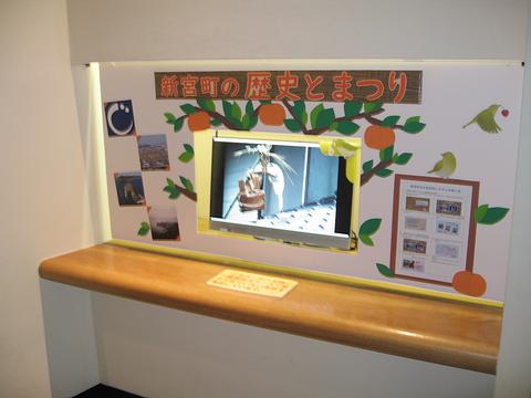 壁に「新宮町の歴史とまつり」と書かれたボードが設置され、ボードの中心部分に祭りの様子がモニターで映し出されている写真