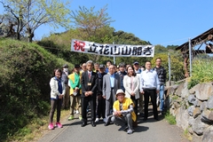 黄色いベストを着た男女や関係者が、立花山山開きと書かれたのぼりの下に集まっている記念写真