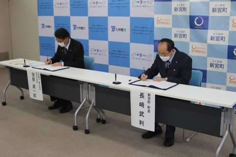 長机に設置された椅子に並んで座り、町長と九州電力の担当者の方が協定書にサインをしている様子を写した写真